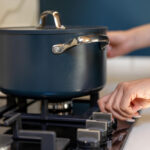 Person adjusting a gas stove knob with a Dutch oven pot cooking on the burner.