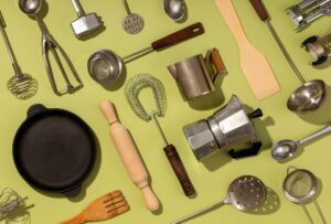Top kitchen essentials including frying pan, whisk, ladle, rolling pin, strainer, and coffee maker on green background