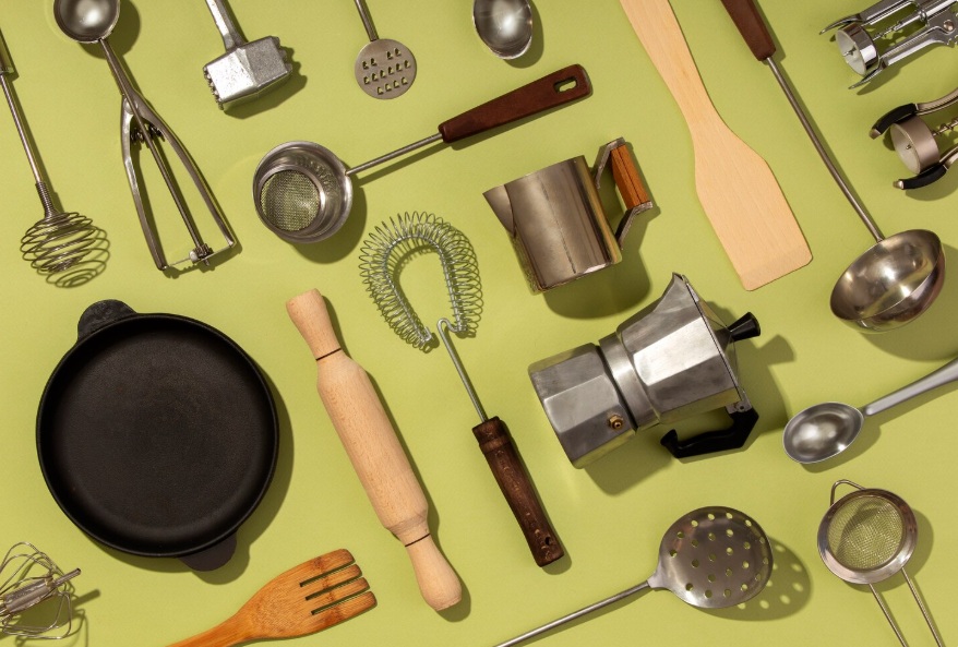Top kitchen essentials including frying pan, whisk, ladle, rolling pin, strainer, and coffee maker on green background