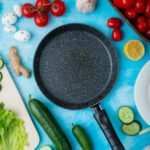 Black non-stick frying pan on a blue surface surrounded by fresh vegetables including tomatoes, cucumbers, garlic, lemon, lettuce, and ginger.