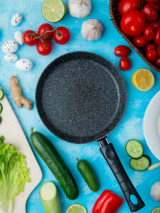 Black non-stick frying pan on a blue surface surrounded by fresh vegetables including tomatoes, cucumbers, garlic, lemon, lettuce, and ginger.