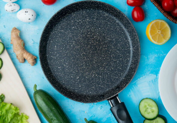 Black non-stick frying pan on a blue surface surrounded by fresh vegetables including tomatoes, cucumbers, garlic, lemon, lettuce, and ginger.