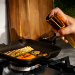 Hand spraying oil onto a cast iron grill pan while seasoning vegetables on a stovetop for cast iron maintenance and nonstick performance.