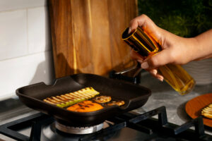 Hand spraying oil onto a cast iron grill pan while seasoning vegetables on a stovetop for cast iron maintenance and nonstick performance.