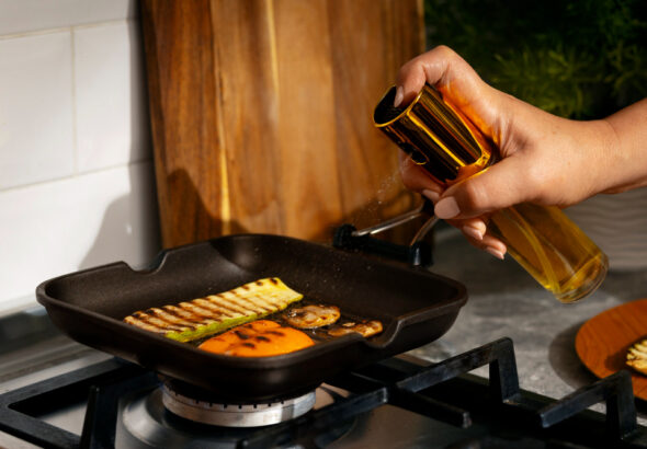Hand spraying oil onto a cast iron grill pan while seasoning vegetables on a stovetop for cast iron maintenance and nonstick performance.