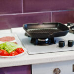Grill pan on a gas stove with fresh burger patties, lettuce, and tomatoes prepared for cooking.