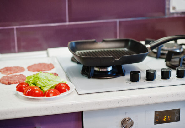 Grill pan on a gas stove with fresh burger patties, lettuce, and tomatoes prepared for cooking.