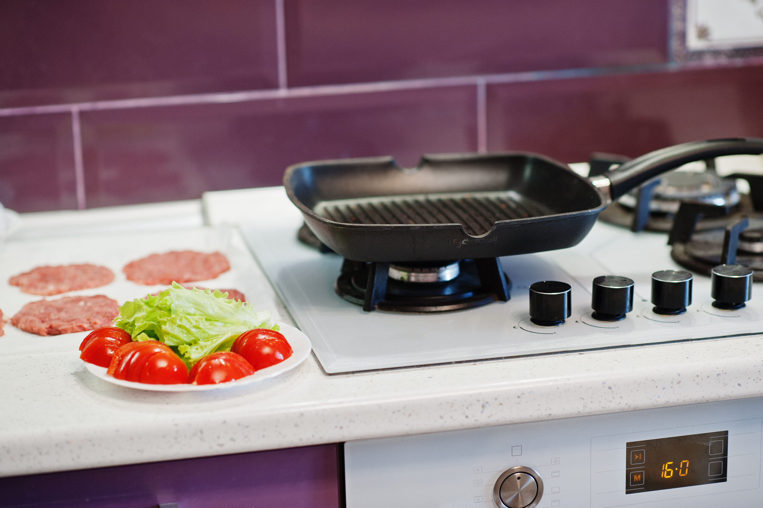 Grill pan on a gas stove with fresh burger patties, lettuce, and tomatoes prepared for cooking.