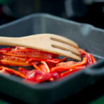 Red bell pepper slices being sautéed in a cast iron skillet with a wooden spatula, highlighting even heat cooking and stovetop meal preparation.