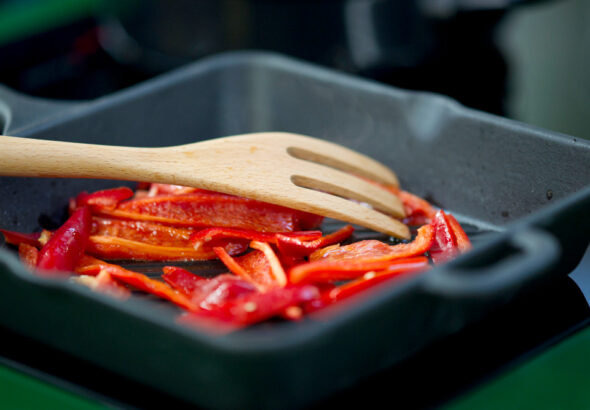 Red bell pepper slices being sautéed in a cast iron skillet with a wooden spatula, highlighting even heat cooking and stovetop meal preparation.
