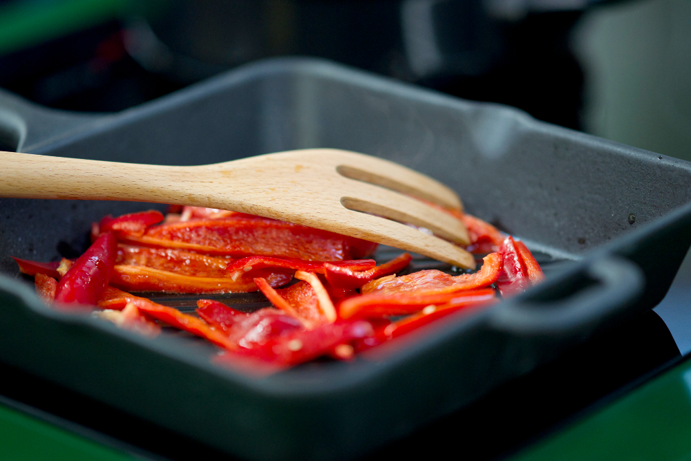 Red bell pepper slices being sautéed in a cast iron skillet with a wooden spatula, highlighting even heat cooking and stovetop meal preparation.
