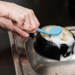 A person scrubs the bottom of a black cooking pot with a blue dish brush covered in soap suds over a kitchen sink.
