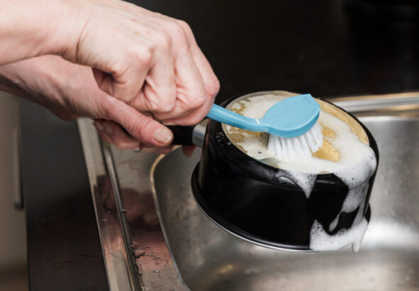 A person scrubs the bottom of a black cooking pot with a blue dish brush covered in soap suds over a kitchen sink.