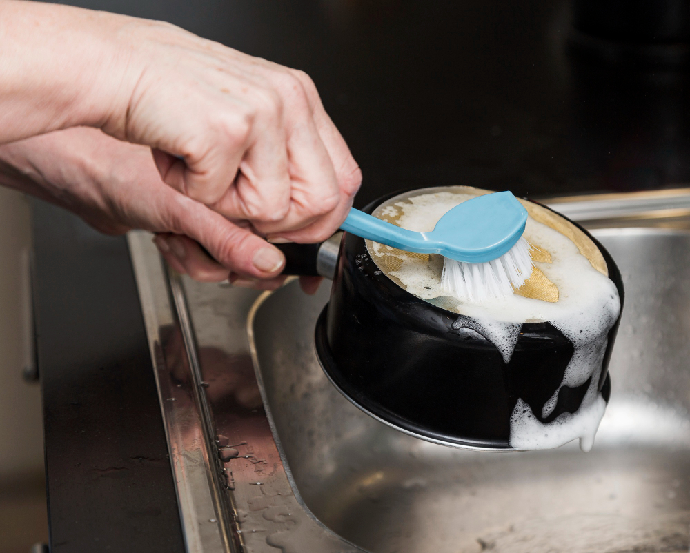 A person scrubs the bottom of a black cooking pot with a blue dish brush covered in soap suds over a kitchen sink.