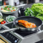 Salmon fillet searing in a frying pan on an induction cooktop, surrounded by fresh vegetables including lettuce, avocado, tomatoes, and herbs.