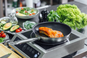 Salmon fillet searing in a frying pan on an induction cooktop, surrounded by fresh vegetables including lettuce, avocado, tomatoes, and herbs.