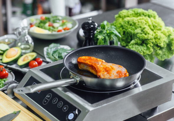 Salmon fillet searing in a frying pan on an induction cooktop, surrounded by fresh vegetables including lettuce, avocado, tomatoes, and herbs.