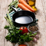 Non-toxic cast iron cookware skillet surrounded by fresh vegetables, herbs, garlic, olive oil, and spices on a rustic wooden table.