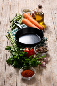 Non-toxic cast iron cookware skillet surrounded by fresh vegetables, herbs, garlic, olive oil, and spices on a rustic wooden table.