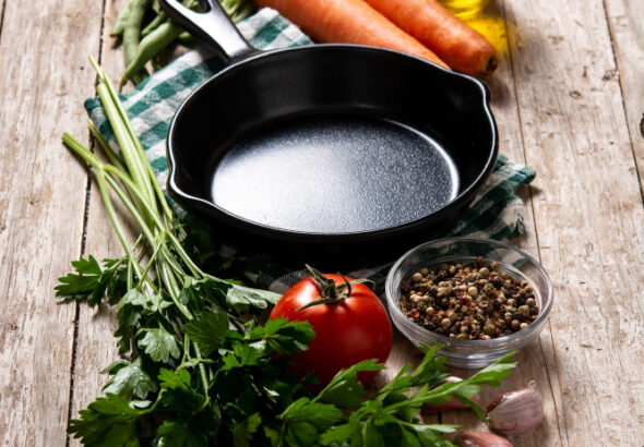 Non-toxic cast iron cookware skillet surrounded by fresh vegetables, herbs, garlic, olive oil, and spices on a rustic wooden table.