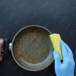 A person wearing a blue glove scrubs a dirty frying pan with a yellow sponge while holding the pan handle against a dark background.