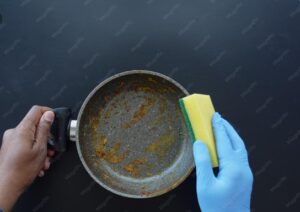 A person wearing a blue glove scrubs a dirty frying pan with a yellow sponge while holding the pan handle against a dark background.